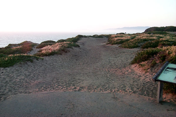 BEGINNING OF IMAGE DESCRIPTION:  THIS IMAGE IS A PHOTOGRAPH OF THE BEGINNING OF THE SUNSET TRAIL NOWADAYS.  IN THE FOREGROUND THE PAVEMENT ENDS AND THE TRAIL CONTINUES ON OVER UNEVEN SAND, WITH ICE PLANT ON EITHER SIDE.  IN THE FAR DISTANCE  IS THE OCEAN, THE ENTRANCE TO SAN FRANCISCO BAY, AND THE MARIN HEADLANDS.  END OF IMAGE DESCRIPTION. 