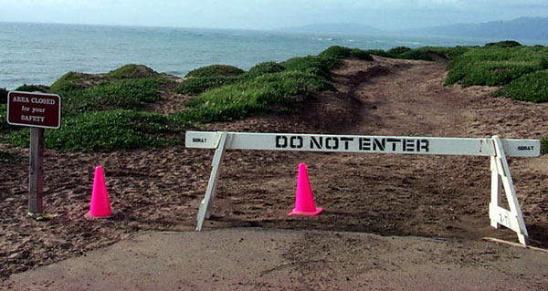 BEGINNING OF IMAGE DESCRIPTION:  THIS IMAGE IS A PHOTOGRAPH OF THE BEGINNING OF THE SUNSET TRAIL DURING THE PERIOD IT WAS CLOSED EARLIER THIS YEAR.  IN THE FOREGROUND THE PAVEMENT ENDS AND THE TRAIL CONTINUES IN THE SAND WHERE THE PAVEMENT HAS BEEN REMOVED.  THERE IS A SIGN THAT SAYS, QUOTE, AREA CLOSED FOR YOUR SAFETY, END QUOTE.  THERE ARE ALSO TWO BRIGHT RED TRAFFIC CONES IN THE PATH.  THERE IS A WHITE SAWHORSE BLOCKING THE TRAIL.  IT HAS VERY LARGE STENCILLED BLACK LETTERS THAT SAY, QUOTE, DO NOT ENTER, END QUOTE.  THE TRAIL CONTINUES INTO THE SAND, WITH ICE PLANT ON EITHER SIDE.  IN THE FAR DISTANCE  IS THE OCEAN, THE ENTRANCE TO SAN FRANCISCO BAY, AND THE MARIN HEADLANDS.  END OF IMAGE DESCRIPTION. 